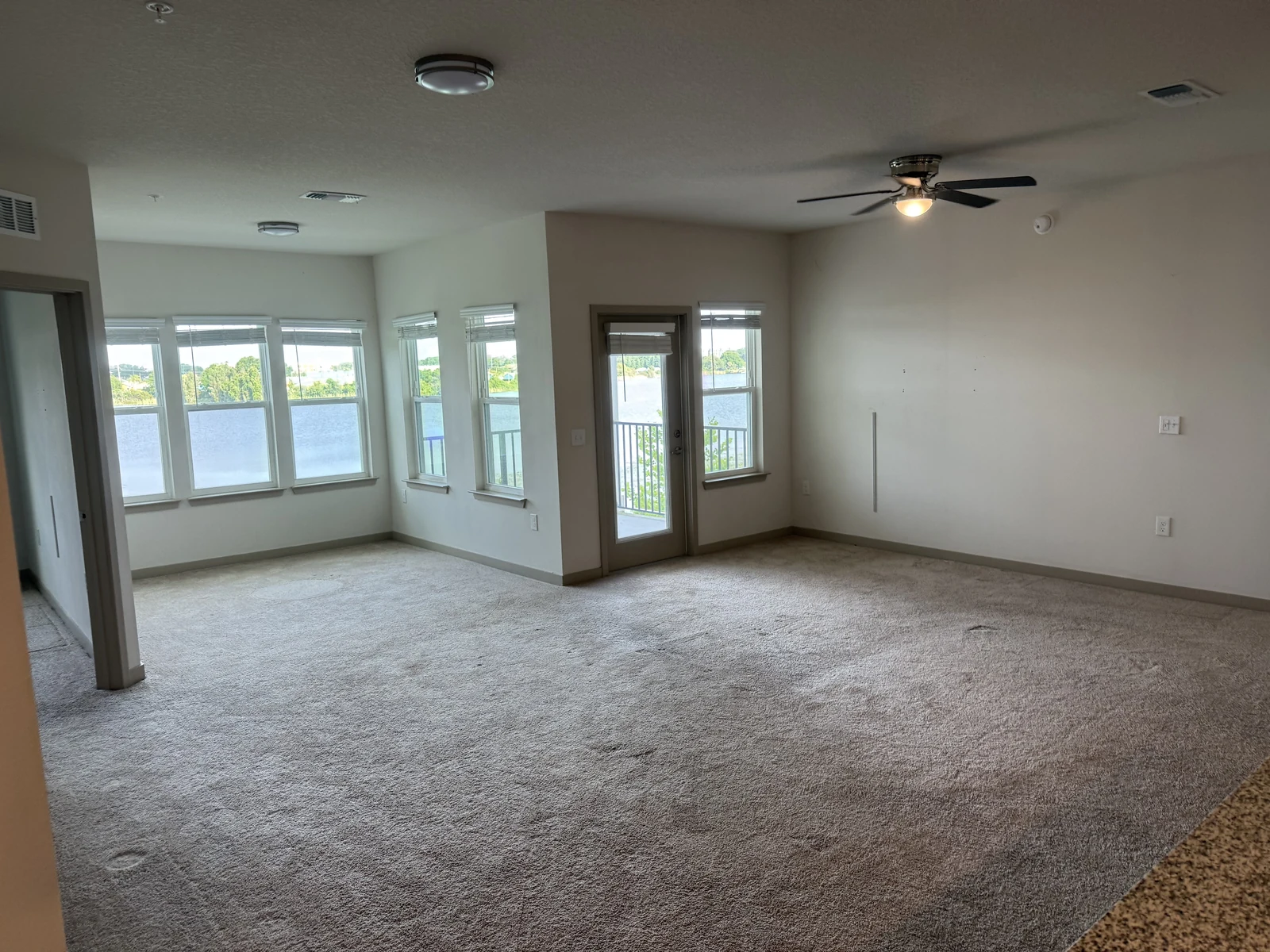 Cleared-out living area with empty carpeted floor, windows and a balcony door.